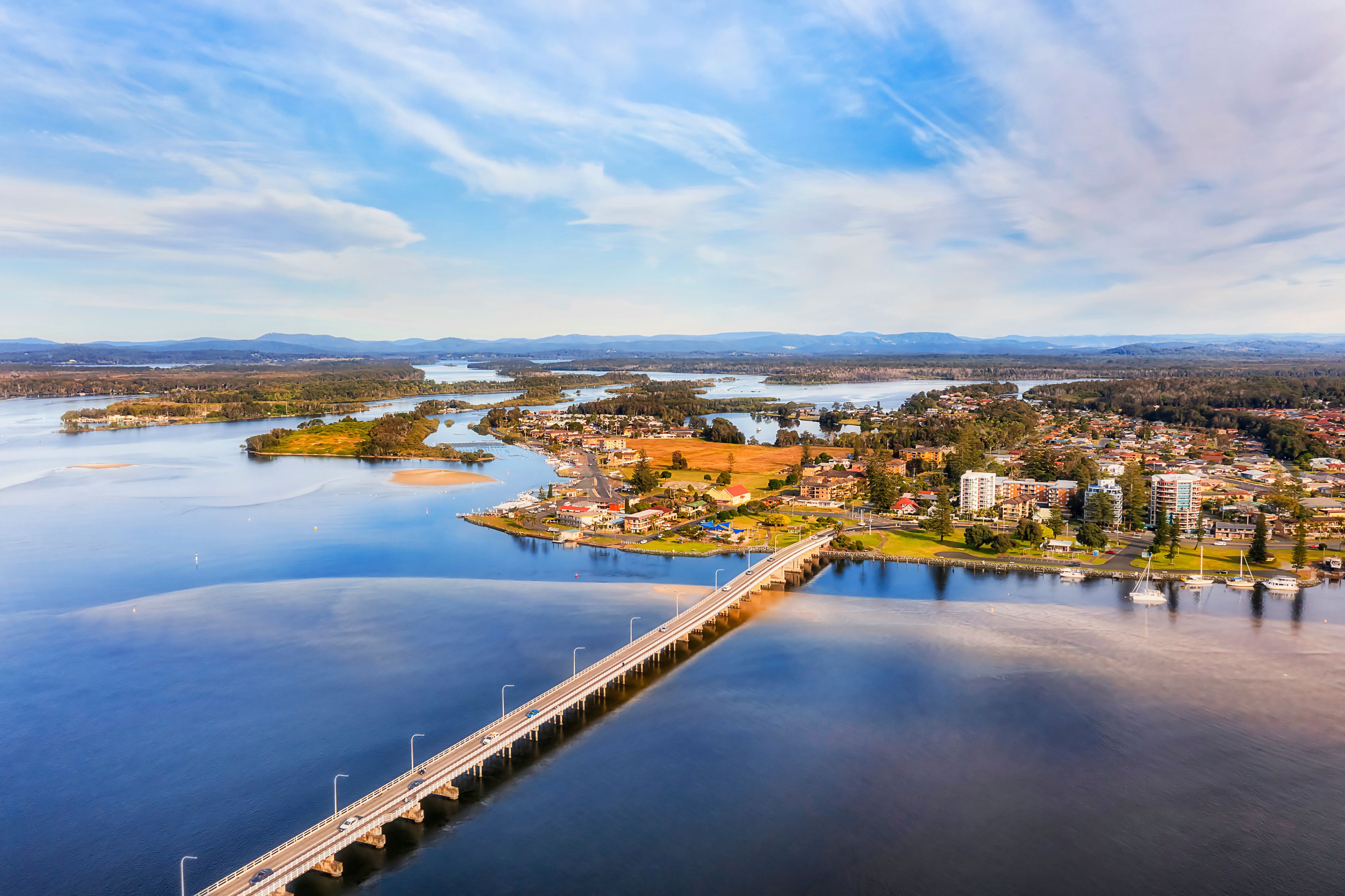 A coastal town with a bridge over a calm river, surrounded by hills and a clear sky.