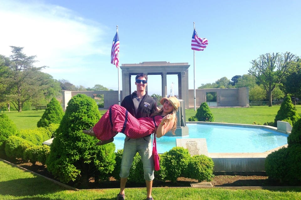 A man carrying a woman in front of a pool with two American flags, surrounded by greenery and a monument.
