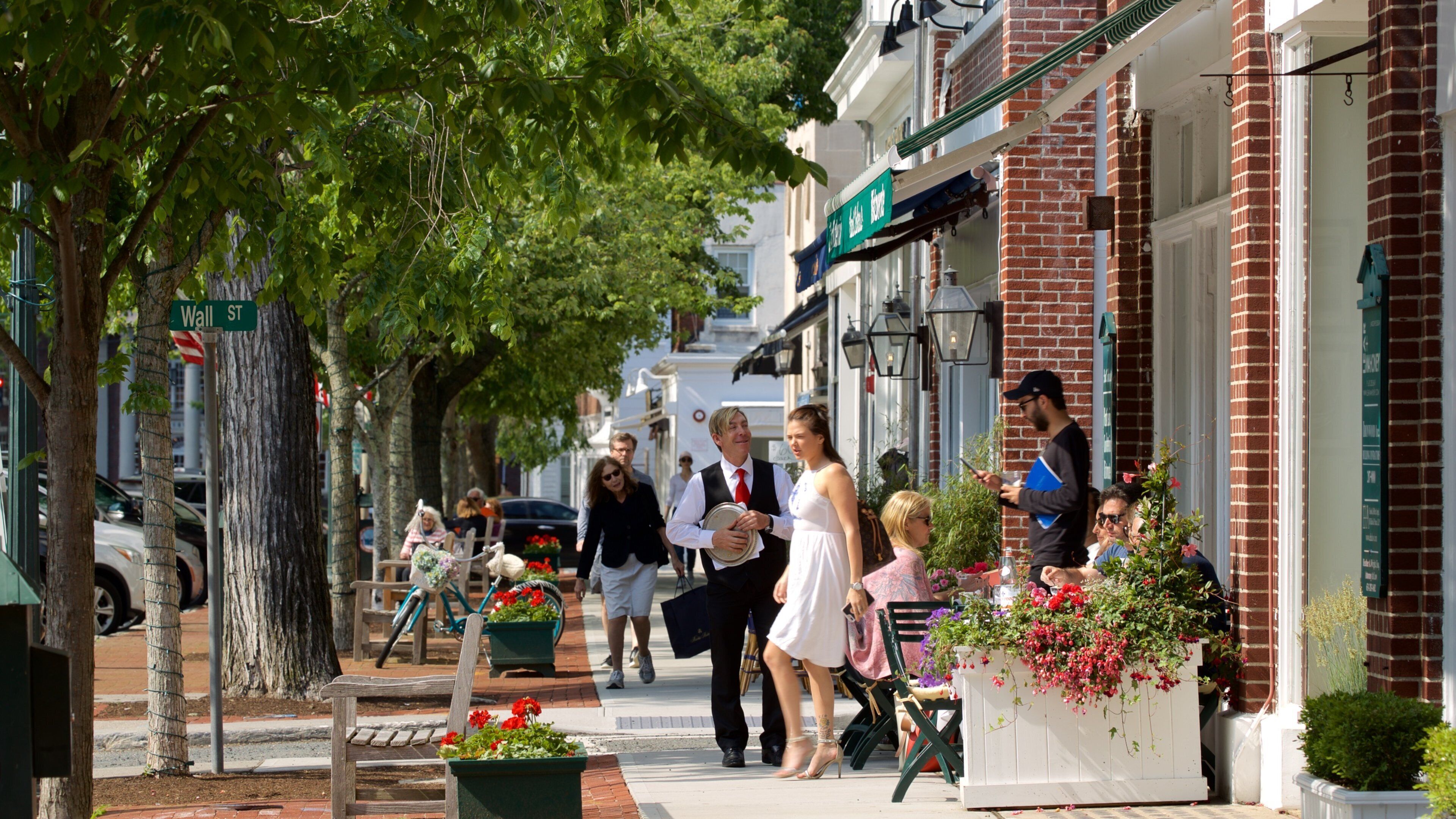 A street lined with brick buildings, a bench, and potted plants with flowers.