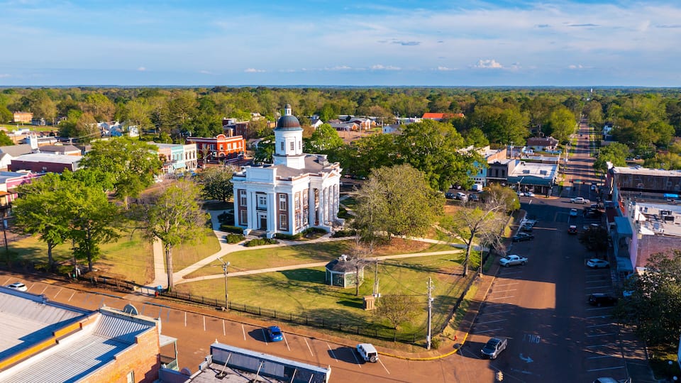 A town with a prominent white church with a dome, surrounded by green spaces and a parking lot.