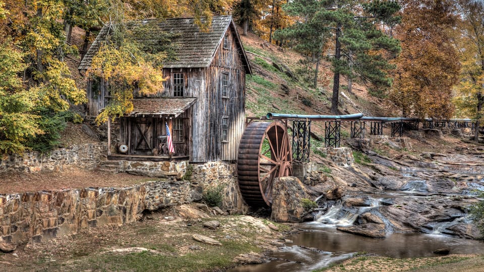A rustic watermill with a wooden structure and a large water wheel, surrounded by autumn foliage and a flowing stream.