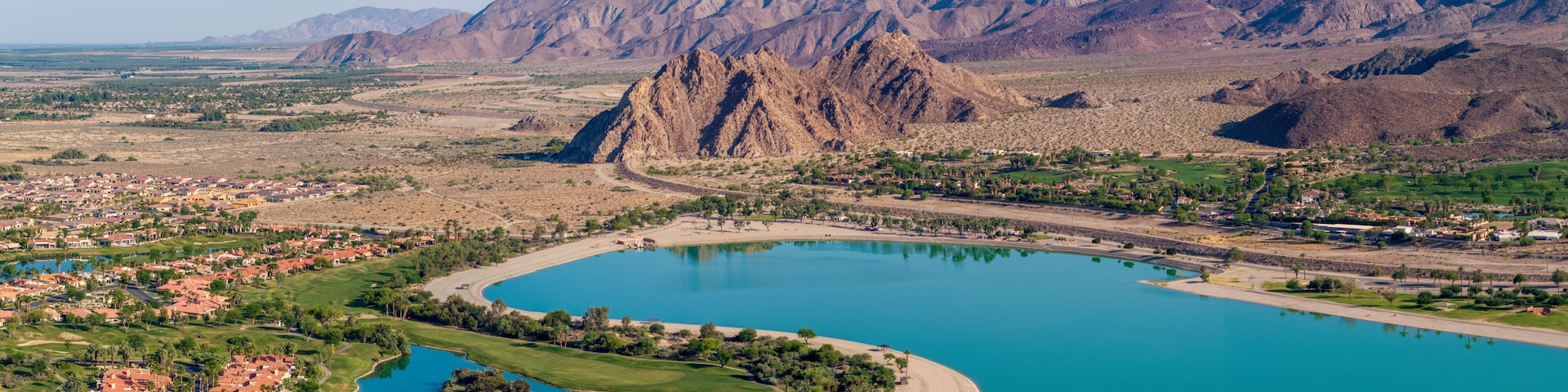 Un pueblo costero con una gran bahía, montañas al fondo y zonas residenciales con casas de techo rojo.