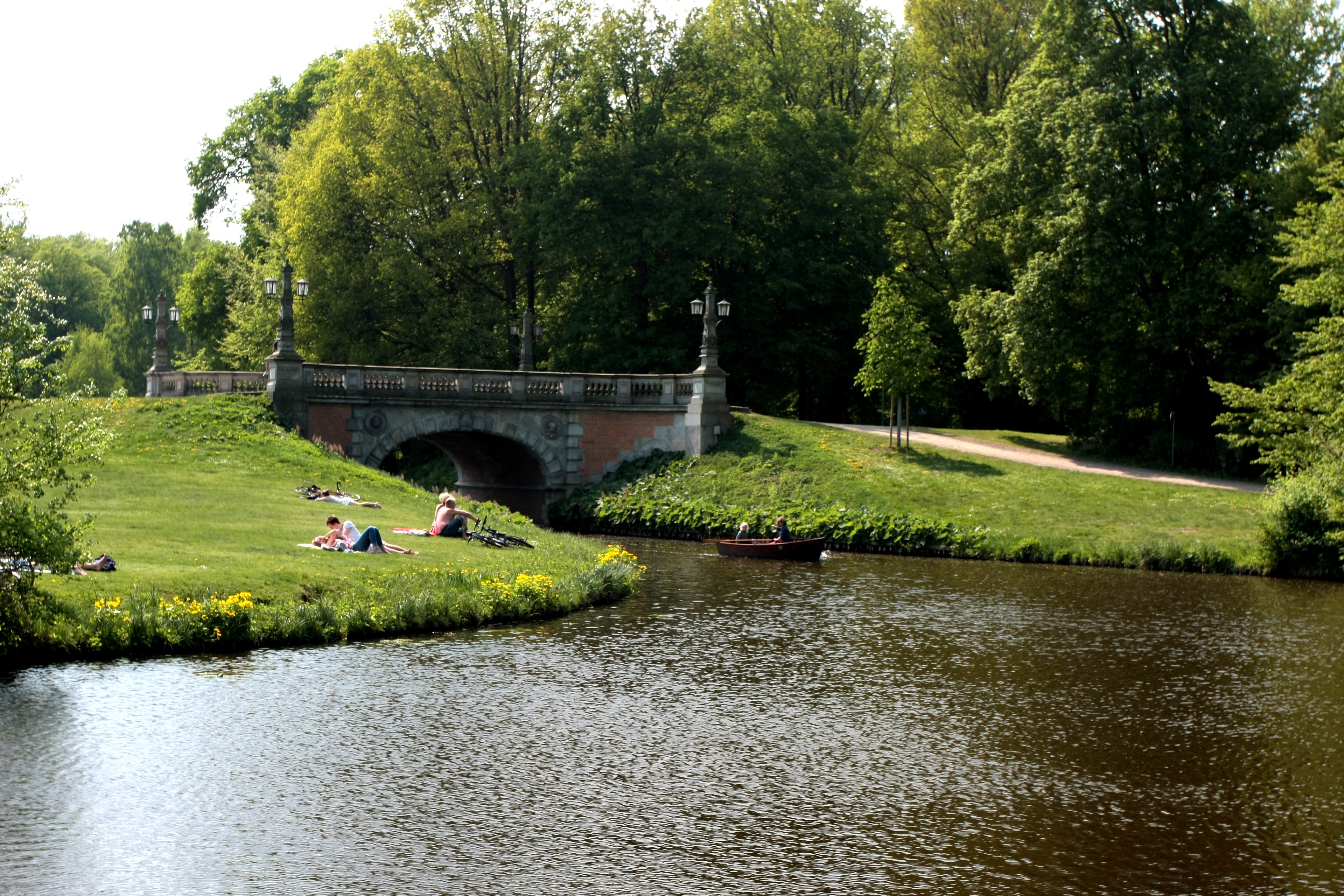 Un parc avec un pont de pierre, une rivière paisible et des gens qui se détendent sur l’herbe.