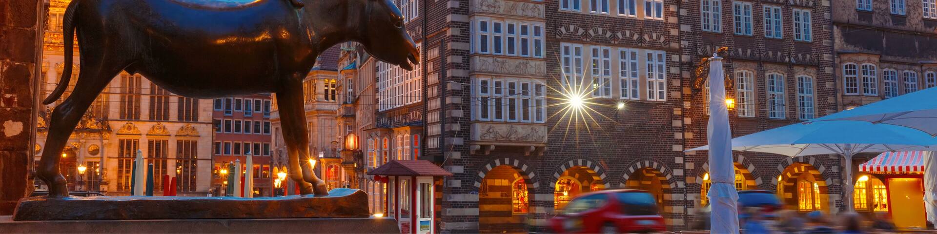 A historic European town square with a prominent statue of a horse and a ram, traditional half-timbered buildings, and a clear evening sky.