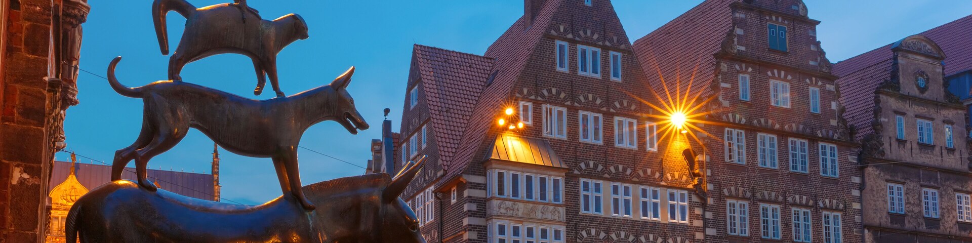 Uma praça histórica europeia com uma estátua imponente de um cavalo e um carneiro, edifícios tradicionais em estilo de enxaimel e um céu claro ao entardecer.