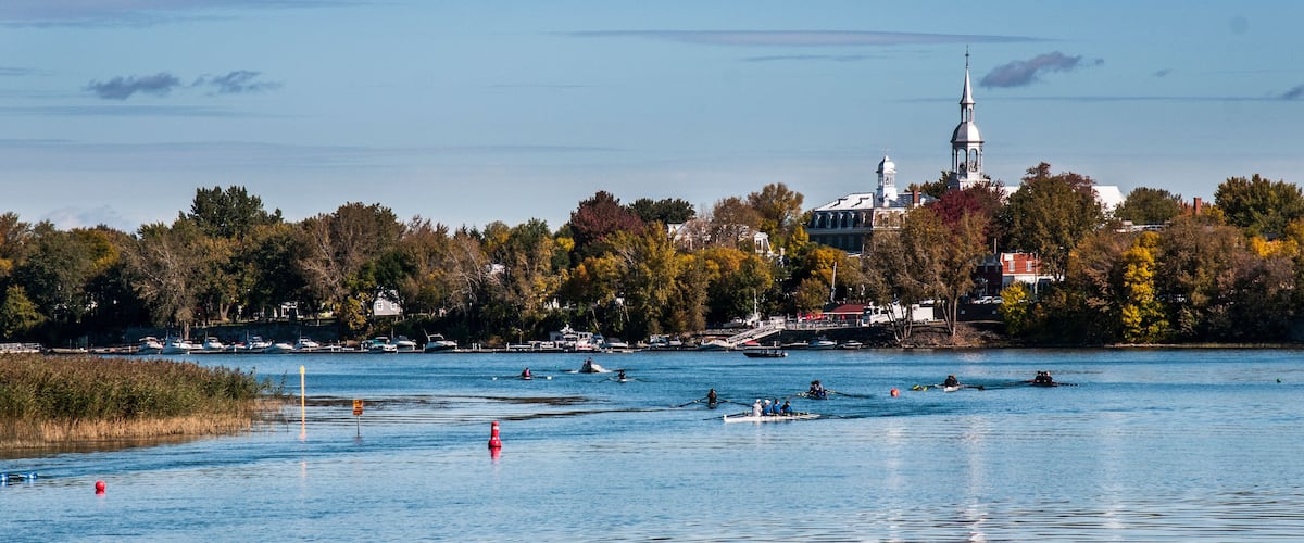 A river with boats, a church with a tall spire, and autumn-colored trees.