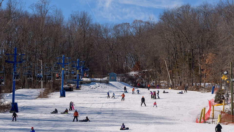 A snowy ski slope with skiers and snowboarders, ski lifts, and a forested area in the background.