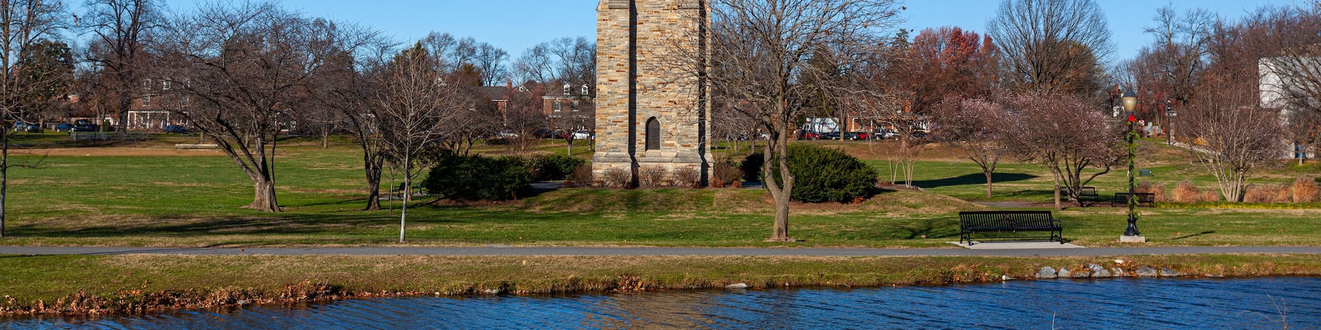 Baker Park, a city park by Carrol Creek with large open meadows, trees, walking and cycling paths and the famous historic tower and carillon. Image shows Frederick cityscape with the river.