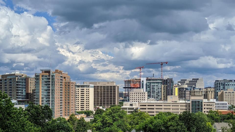 A city skyline with multiple high-rise buildings, construction cranes, and a cloudy sky.