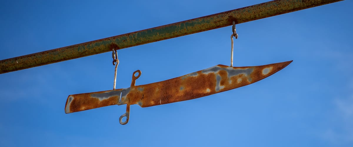 A rusty knife blade suspended by a metal bar against a clear blue sky.