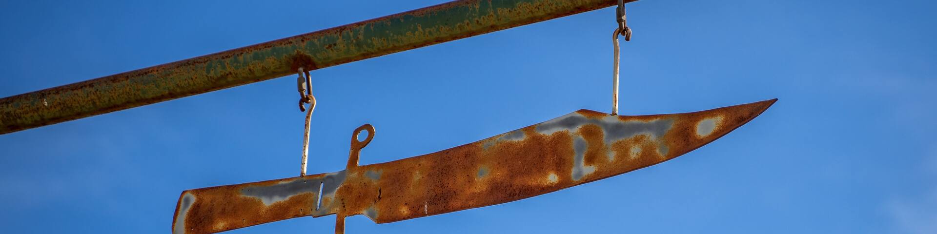 A rusty knife blade suspended by a metal bar against a clear blue sky.