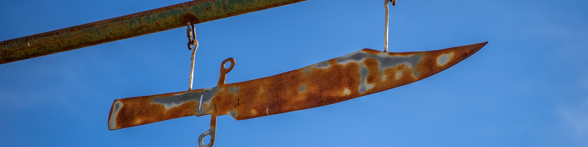 Una hoja de cuchillo oxidada, suspendida por una barra de metal contra un cielo azul despejado.