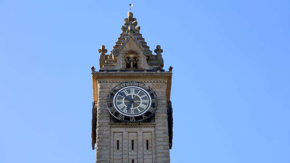 A clock tower with a large Roman numeral clock, a pointed roof, and a weather vane.