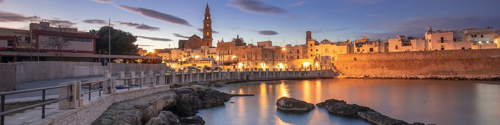 Un paisaje urbano costero al atardecer con una torre destacada, aguas tranquilas y un muro de piedra.