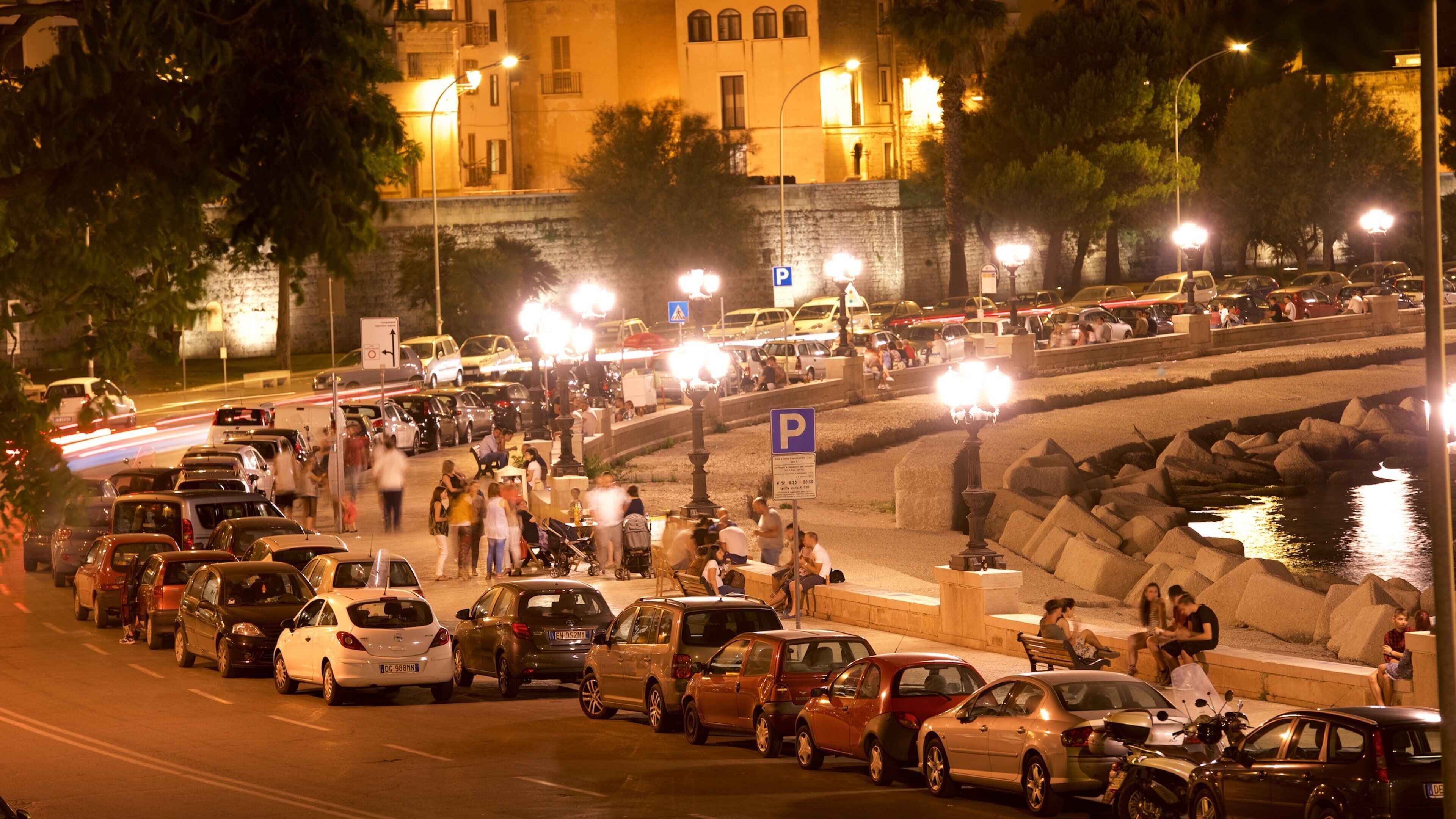 A busy street at night with parked cars, people walking, and a body of water.