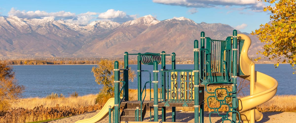A playground with a slide and climbing structures, set against a backdrop of mountains and a lake.