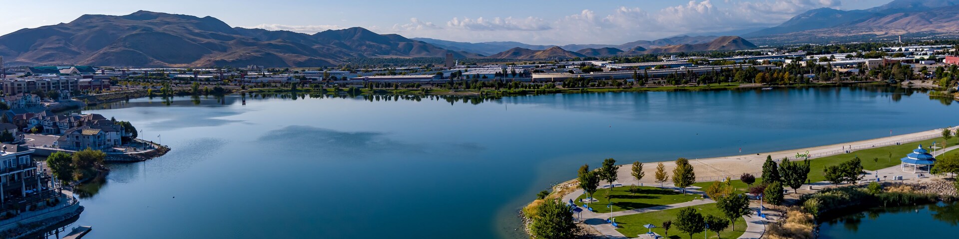 Um lago tranquilo com um píer, um parque com um gazebo e montanhas ao fundo.