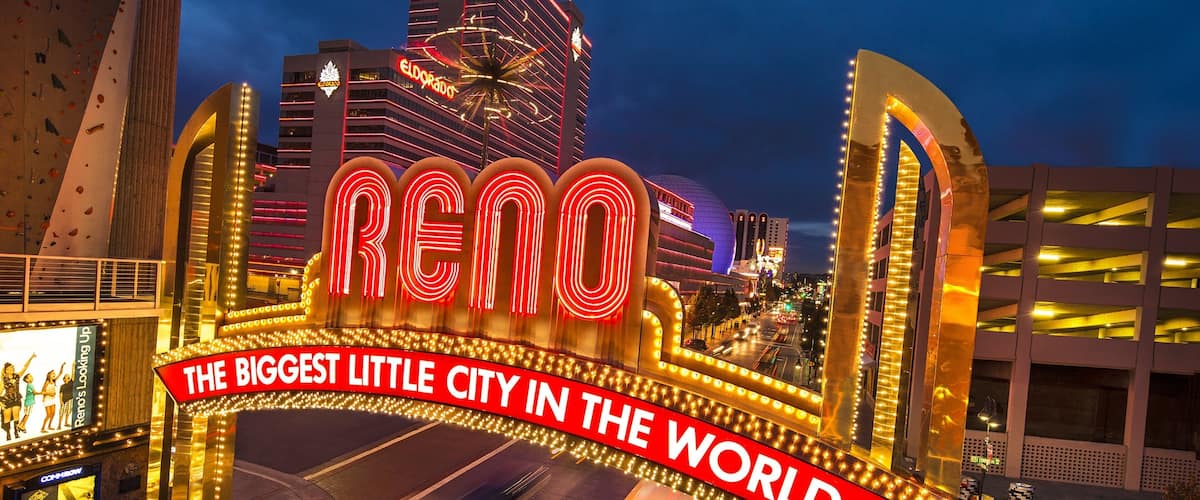 A brightly lit sign with "RENO" and "THE BIGGEST LITTLE CITY IN THE WORLD" text, surrounded by illuminated arches and a cityscape at dusk.