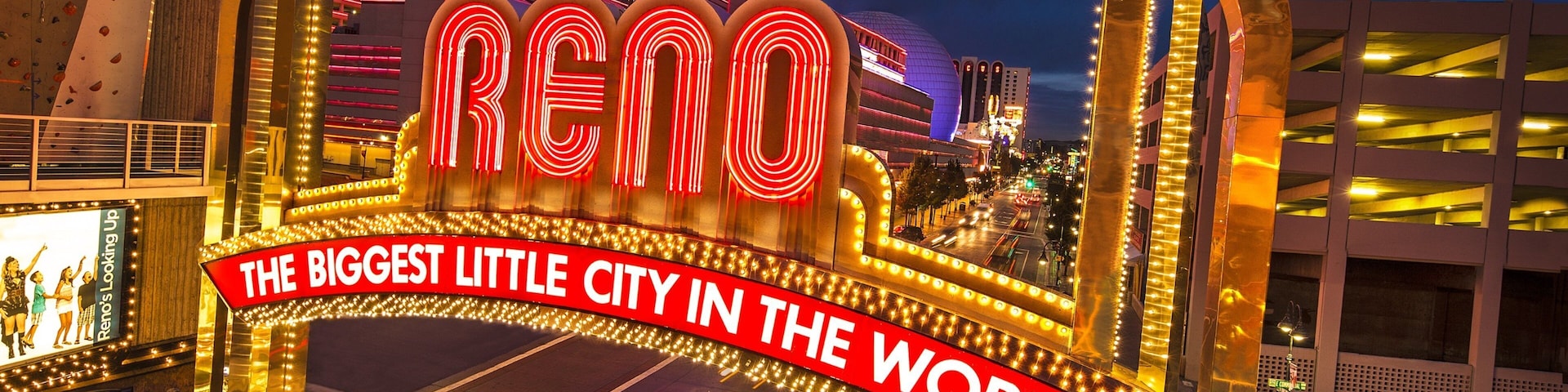 A brightly lit sign with "RENO" and "THE BIGGEST LITTLE CITY IN THE WORLD" text, surrounded by illuminated arches and a cityscape at dusk.