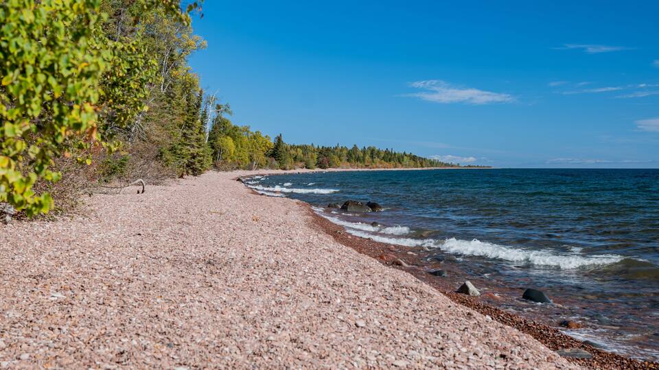 A pebble beach with a forested shoreline and calm sea.