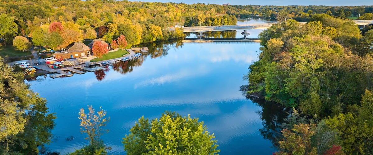 A calm river with a bridge, surrounded by autumn-colored trees and a dock with boats.