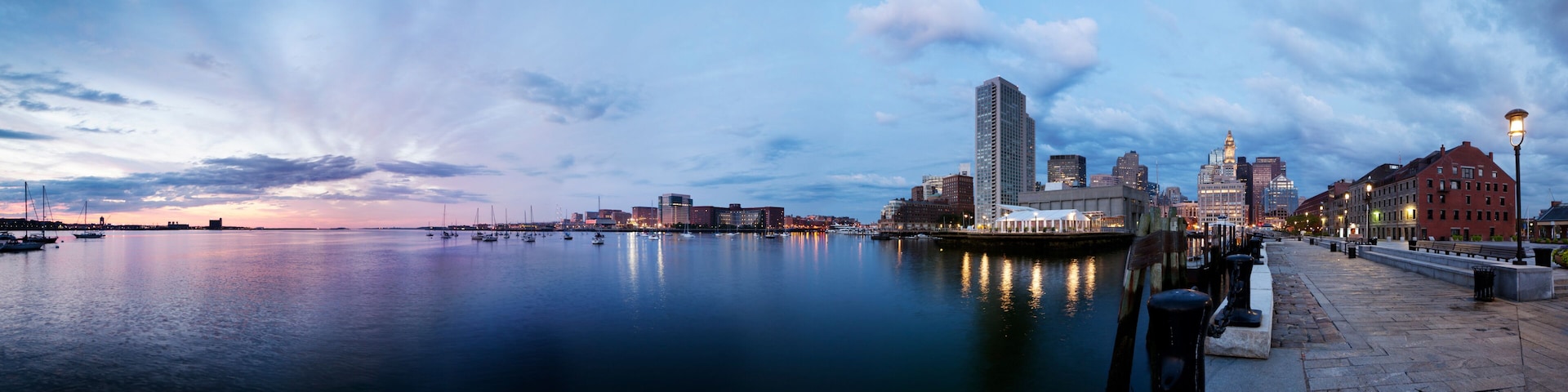 A waterfront cityscape at dusk with calm water reflecting buildings and a clear sky.