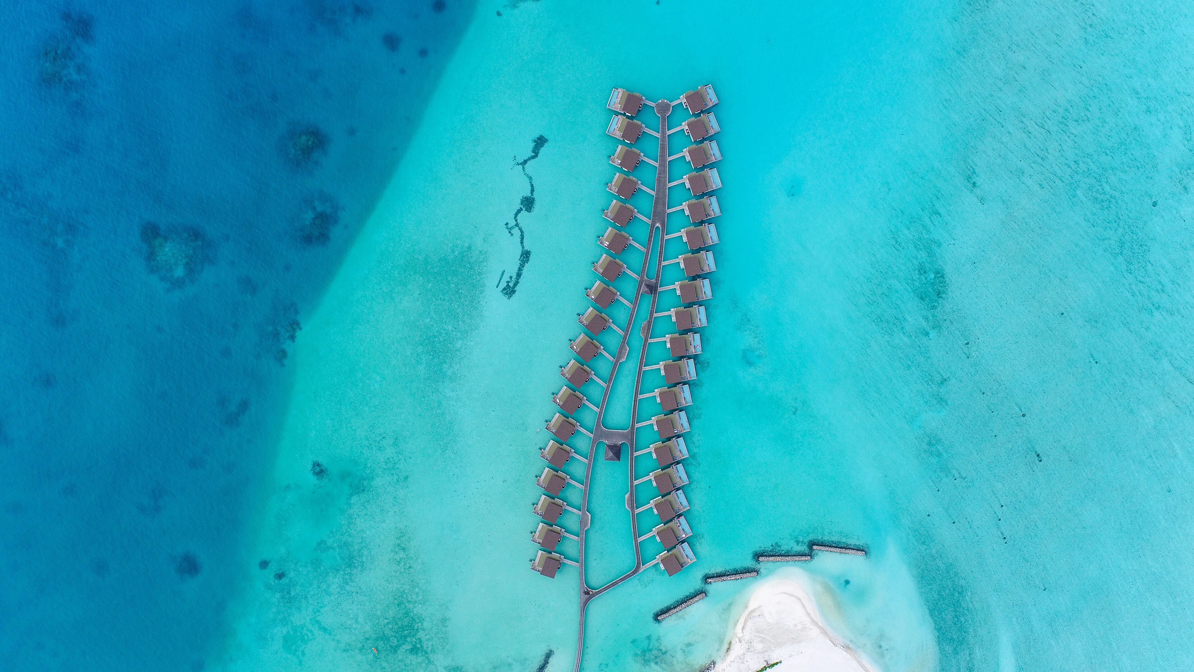 An aerial view of a resort with a long pier extending into turquoise waters, surrounded by clear skies.