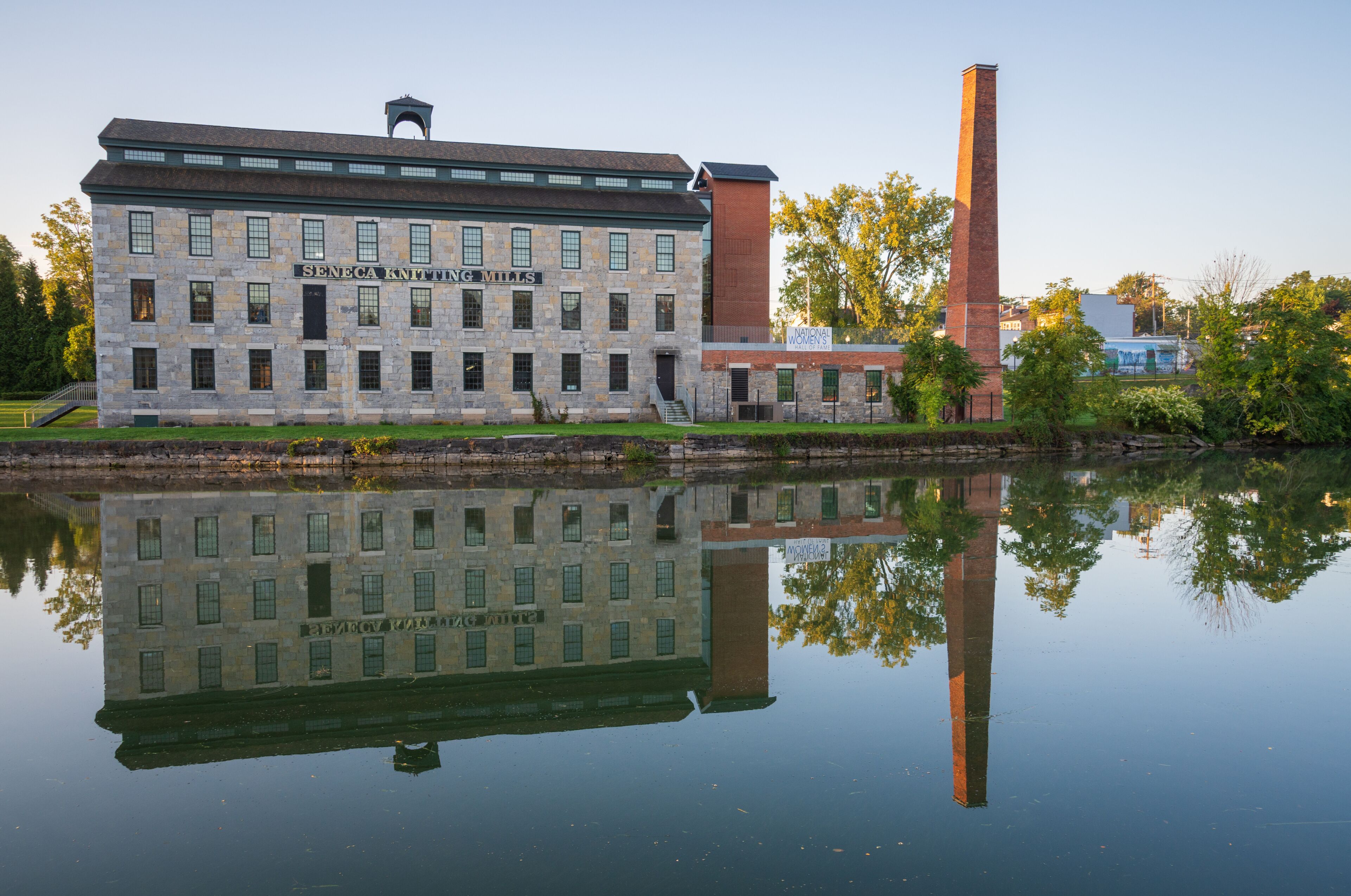 A historic building with a sign reading 'Seneca Knitting Mills' and a tall chimney, reflecting in a calm body of water.