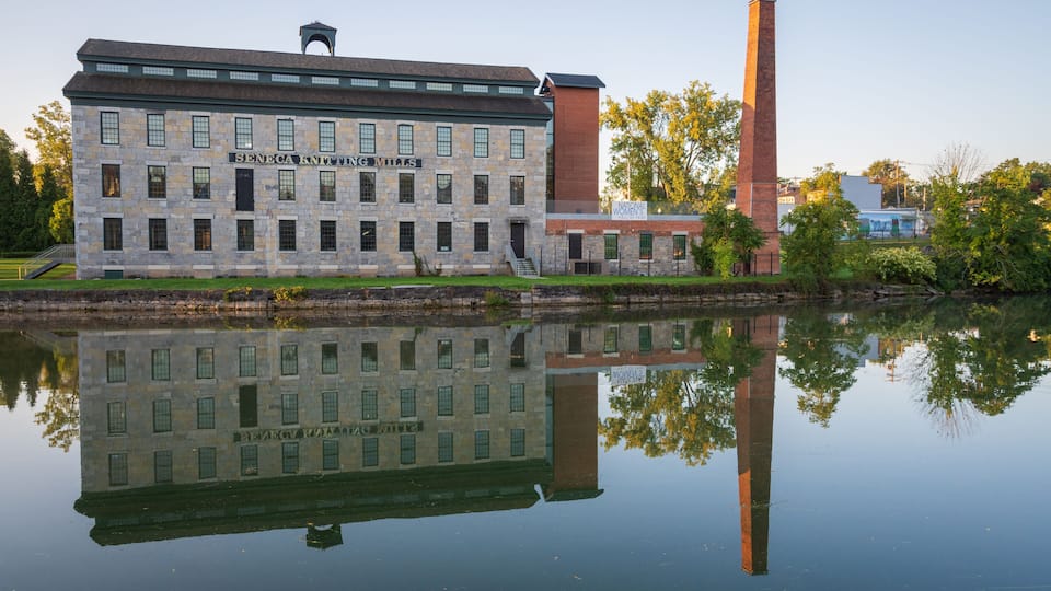 A historic building with a sign reading 'Seneca Knitting Mills' and a tall chimney, reflecting in a calm body of water.