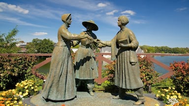 Een bronzen beeld van drie figuren in traditionele kledij, omringd door kleurrijke bloemen en een brug op de achtergrond.