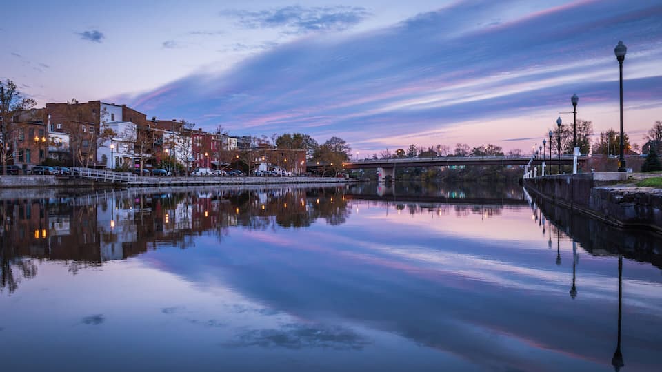 A calm river reflecting buildings and a bridge at dusk.