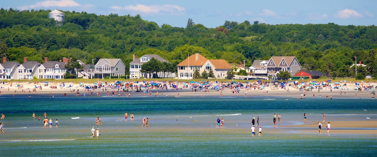 A crowded beach with people sunbathing and swimming, surrounded by houses and lush green hills.