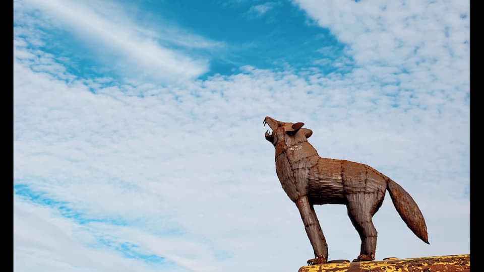 A rusted metal wolf sculpture on a yellow structure against a blue sky with clouds.