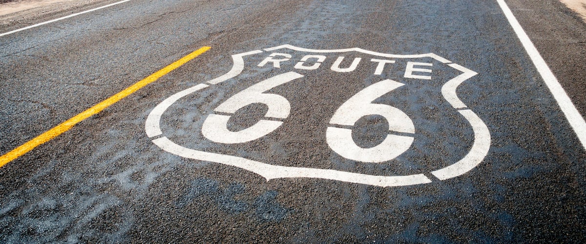 Una carretera con la Ruta 66 pintada sobre el asfalto, rodeada de vegetación seca y líneas eléctricas a lo lejos.
