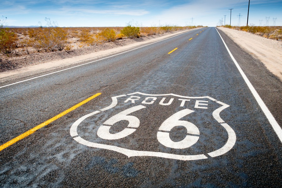 Una carretera con la Ruta 66 pintada sobre el asfalto, rodeada de vegetación seca y líneas eléctricas a lo lejos.