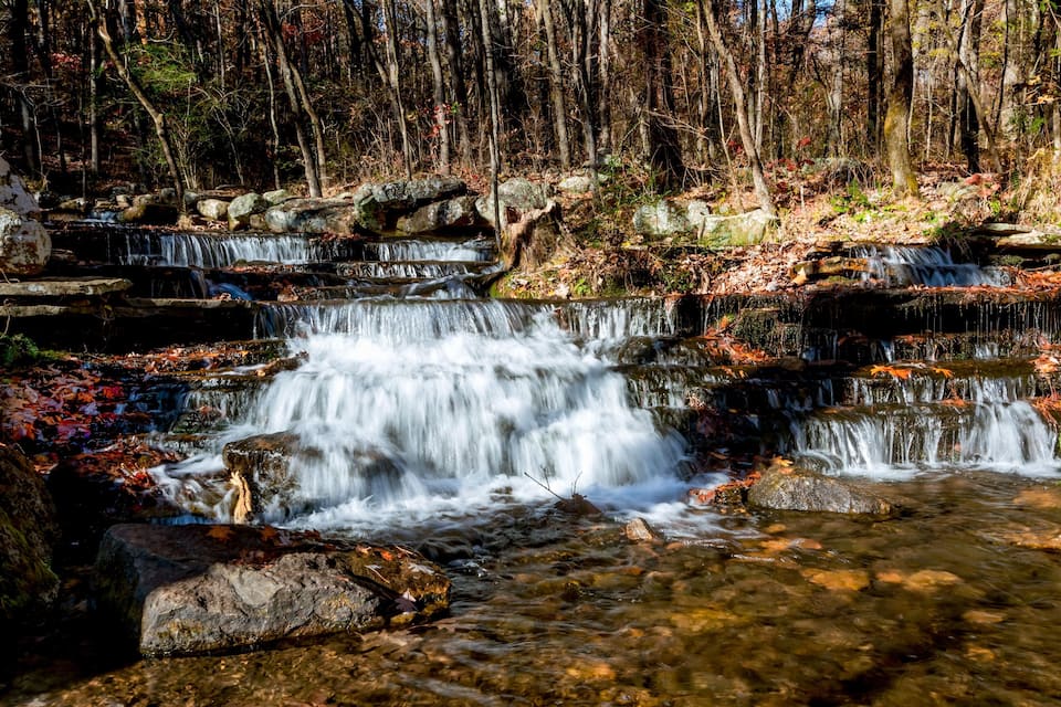 A multi-tiered waterfall in a forested area with rocks and autumn leaves.