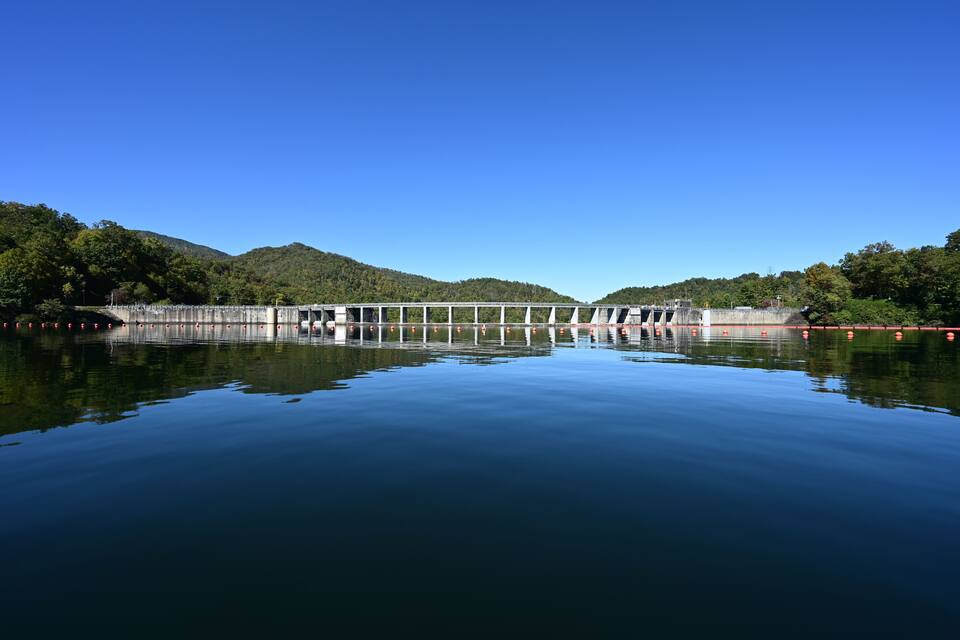 A dam with a bridge over a calm river, surrounded by green hills.