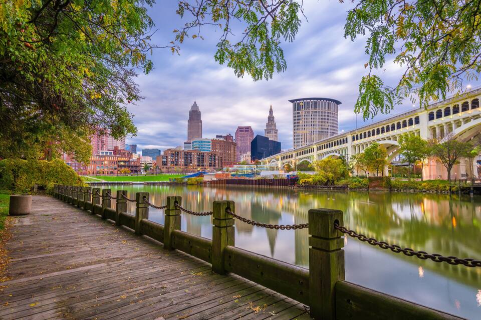 A wooden boardwalk along a river with a city skyline in the background.