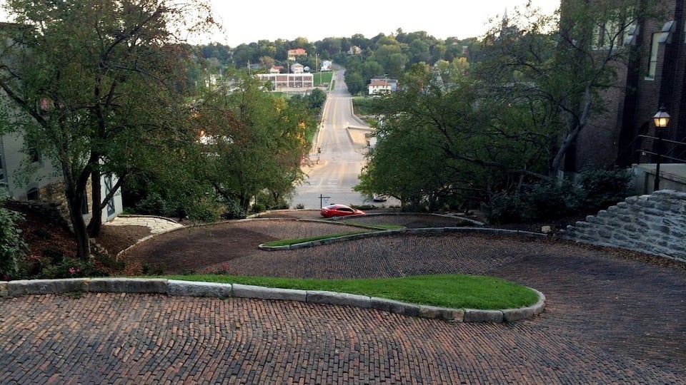 A paved courtyard with a brick pathway, trees, and a distant view of a road and buildings.