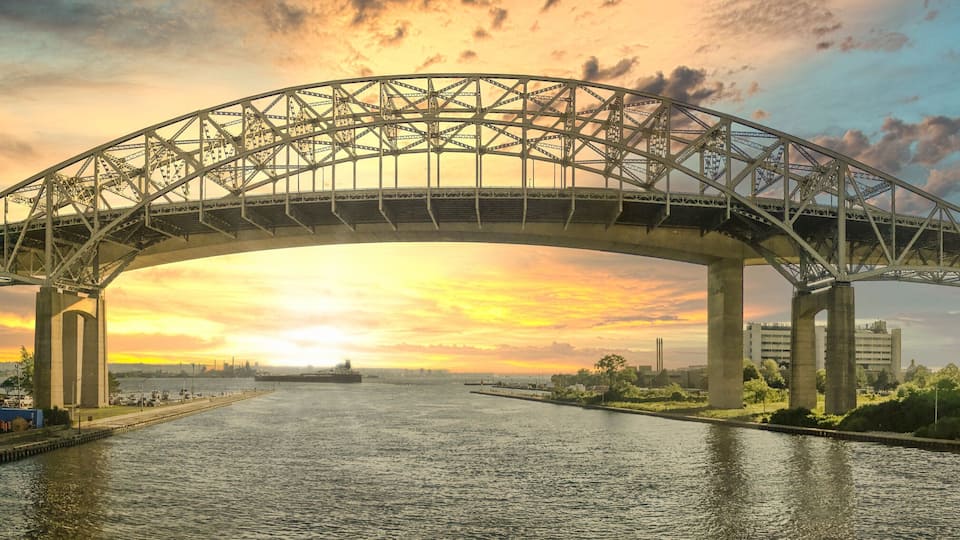 A large steel bridge spanning a river with a sunset in the background.