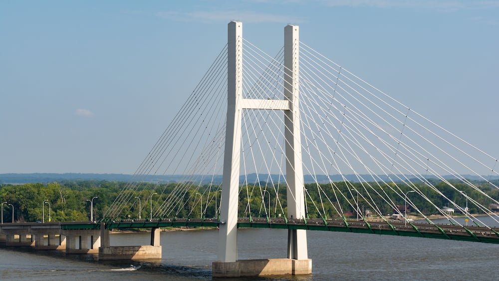 A large white cable-stayed bridge over a river with green supports and a forested landscape in the background.