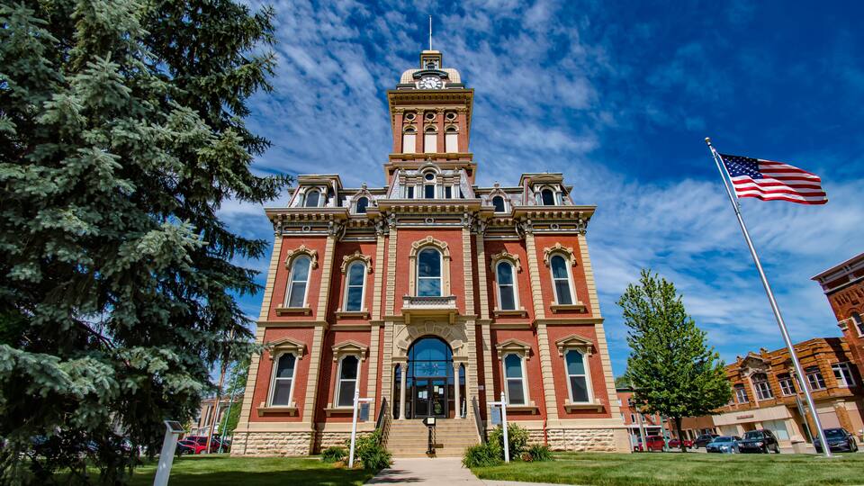 A historic red brick building with a clock tower, surrounded by green trees and a clear blue sky.