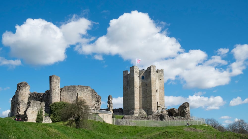 A historic stone castle with a flag on top, surrounded by lush green hills and a clear blue sky.