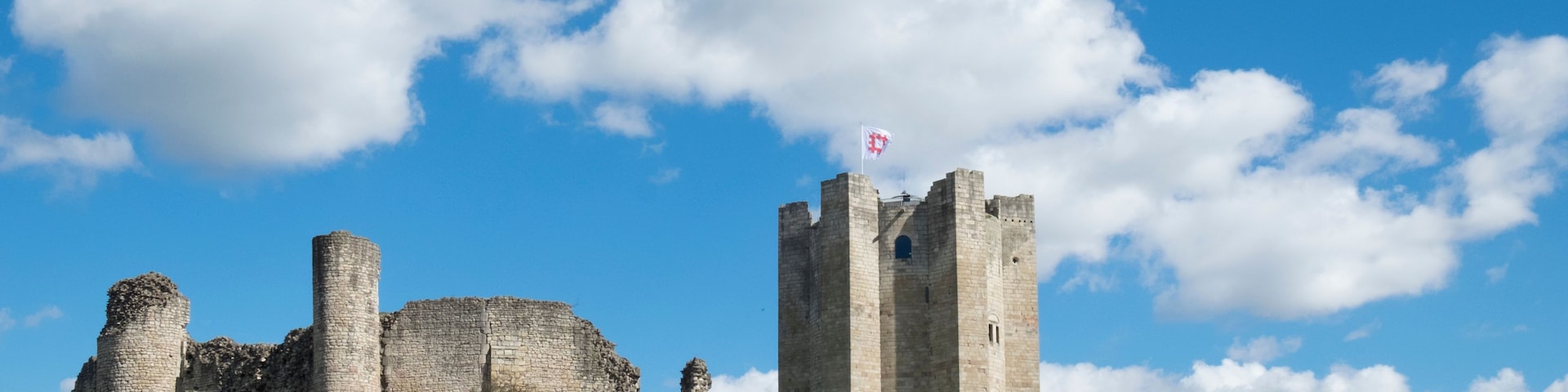 Un castillo de piedra histórico con una bandera en la cima, rodeado de colinas verdes exuberantes y un cielo azul despejado.