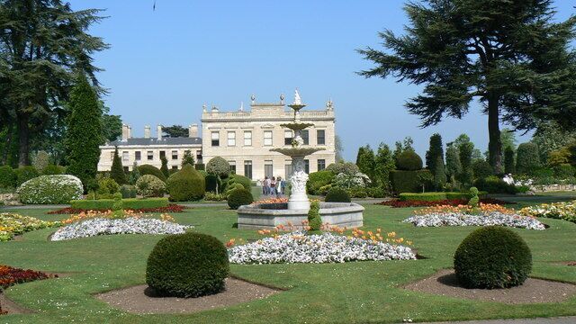 Un jardín formal con una fuente, setos cuidados y un edificio grande al fondo.