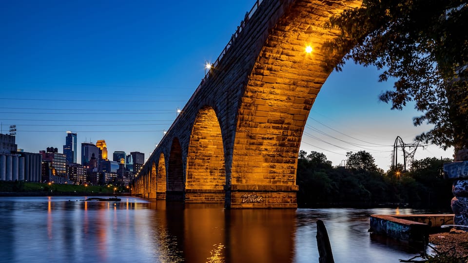 A stone bridge over a river at dusk with city buildings in the background.