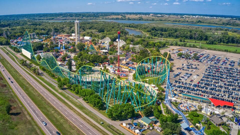 An aerial view of a theme park with roller coasters, a parking lot, and a river in the background.