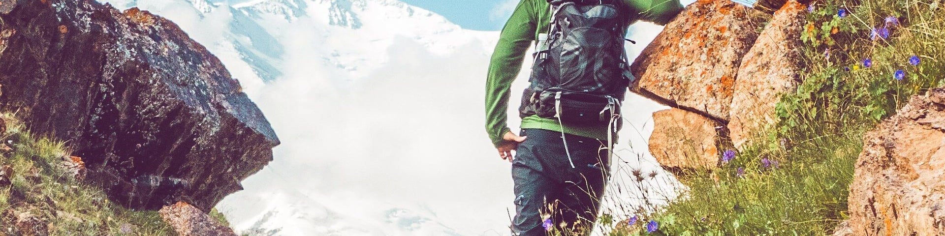 A hiker on a rocky mountain trail with snow-capped peaks in the background.