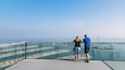 Una pareja de pie en un balcón con vistas a una ciudad costera bajo un cielo despejado.