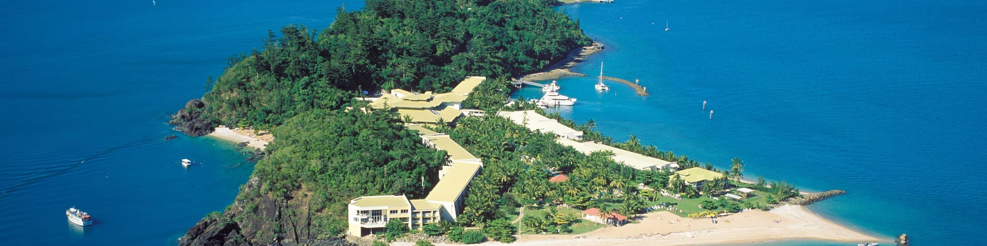 An aerial view of a tropical island with a large building, clear blue waters, and distant mountains.